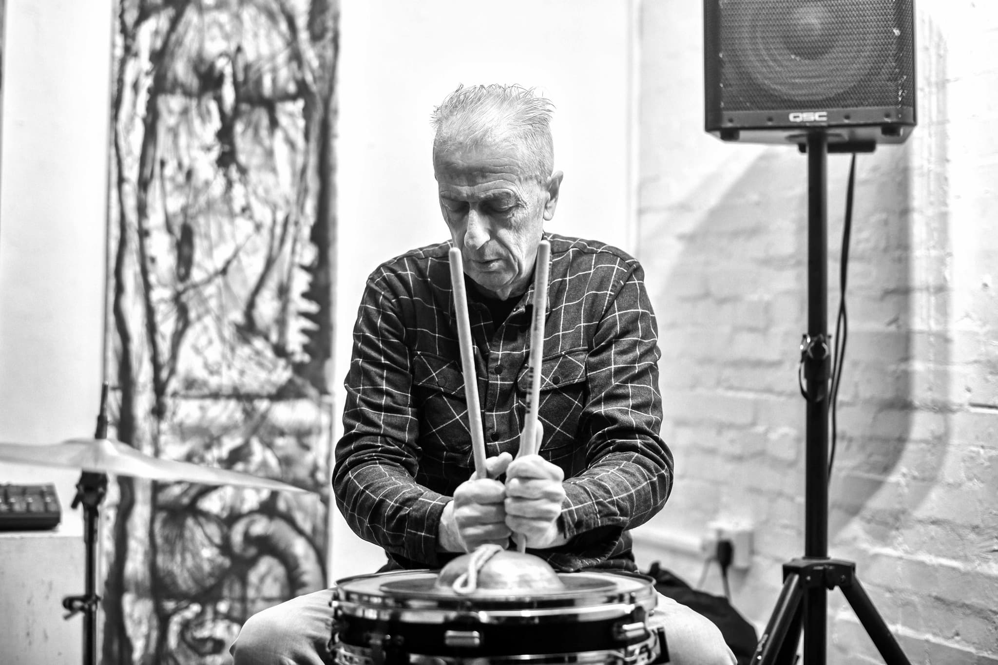 A person is playing percussion with drumsticks, seated in a room with a speaker and abstract artwork in the background.