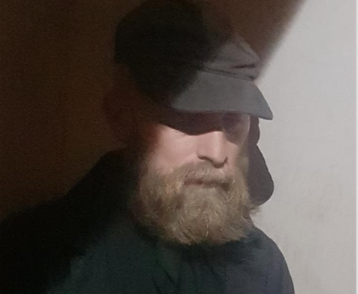 A bearded man wearing a dark cap and coat stands in dim lighting, casting a shadow on part of his face.