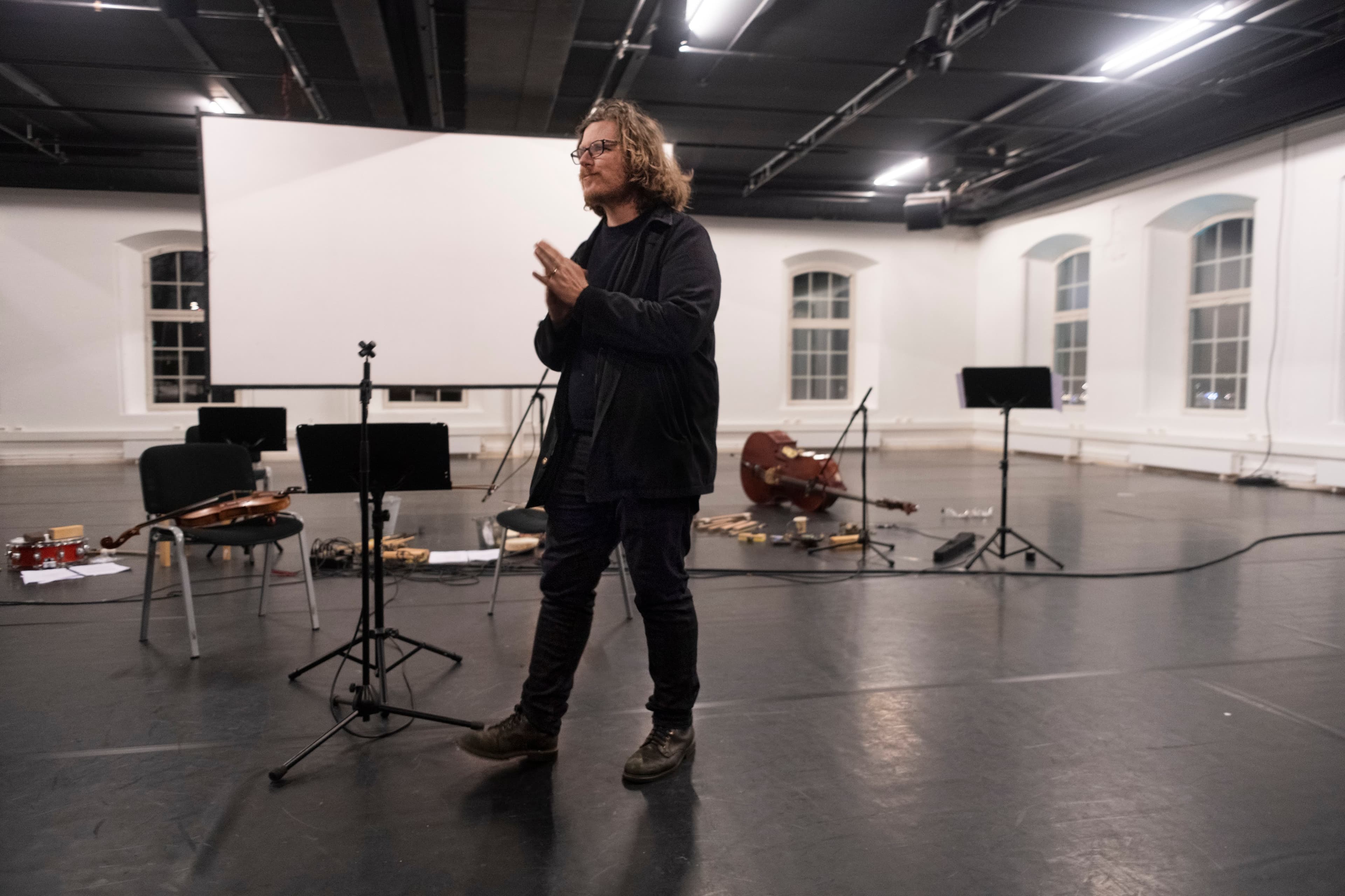 A person stands in a spacious room with musical instruments on the floor and chairs around.