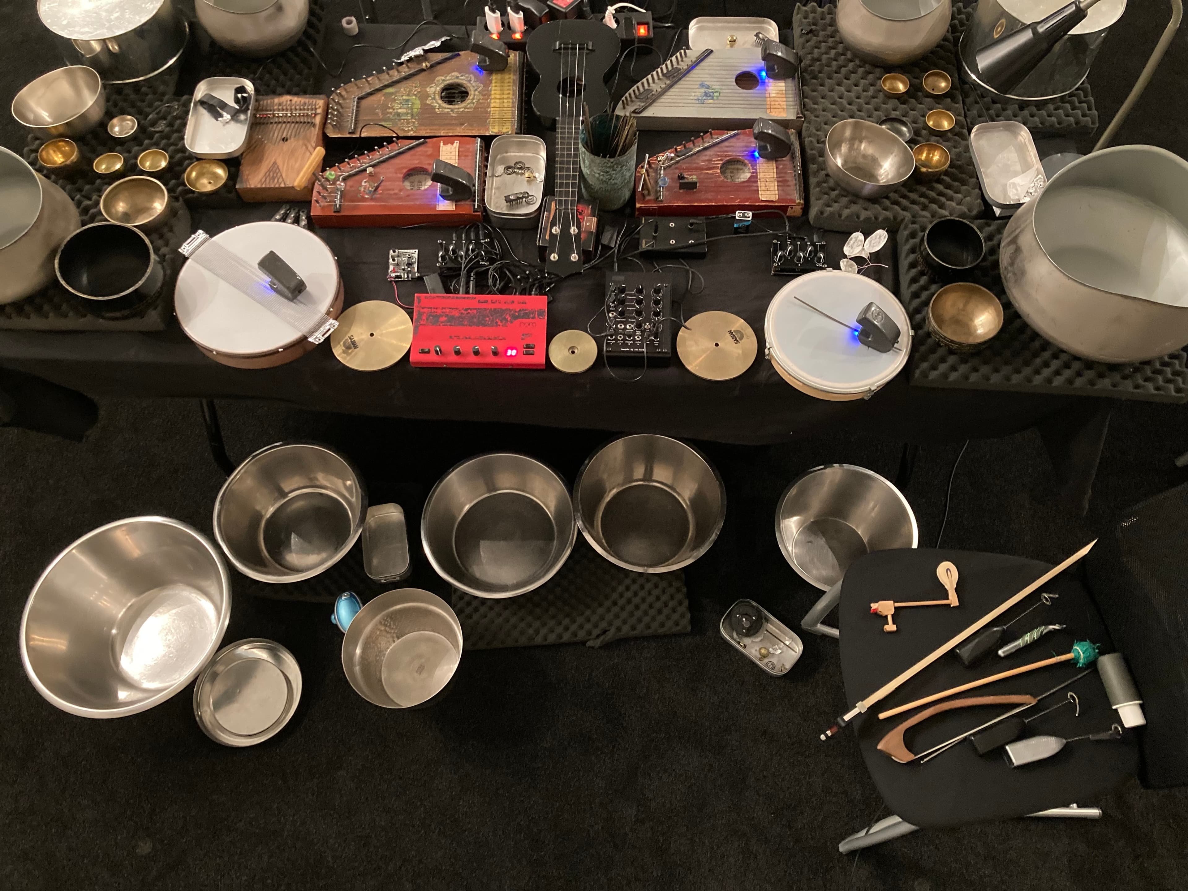A collection of musical instruments and equipment, including metal bowls, zithers, and electronic devices, arranged on a black surface.