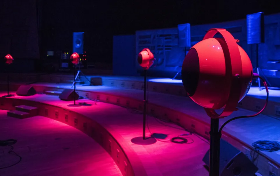 A dimly lit room features multiple spherical speakers on stands, illuminated by red and blue lighting.